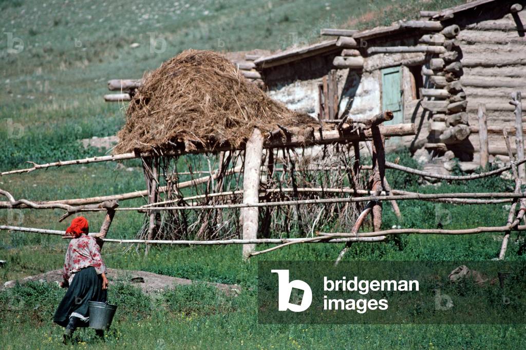 Kazakh woman carrying water buckets from farmhouse built out of wood and mud in hills North of Urumqi, Xinjiang Province, China (photo)