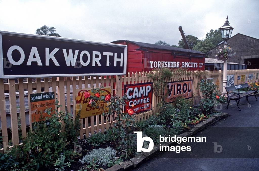 Oakworth station with milk churns, old luggage cart, enamel 1950s advertising signs on the heritage Keighley and Worth Valley Railway, England, UK, 1989 (photo)