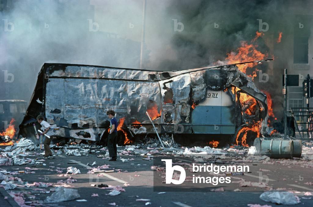 Children playing amongst debris from Hijacked Burning Vehicles after Riots in West Belfast during The Troubles, Northern Ireland, 1976 (photo)
