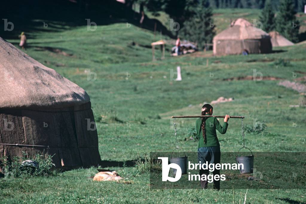 Kazakh woman carrying water with pole and two buckets on shoulder in Yurt settlement in hills North of Urumqi, Xinjiang Province, China (photo)