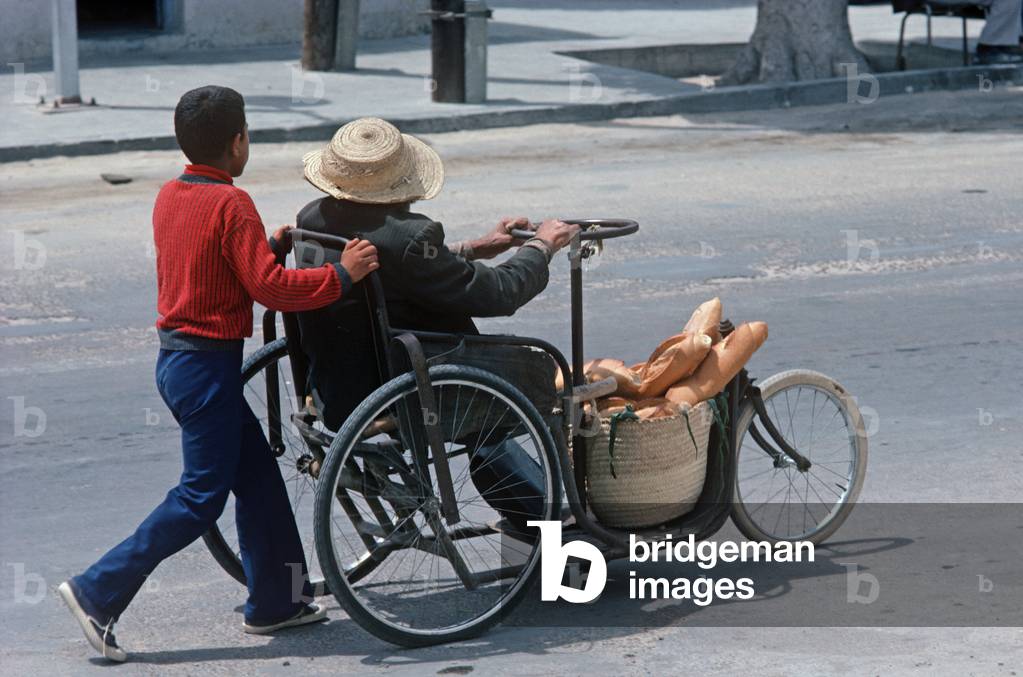 Child pushing disabled man in wheelchair with basket of loaves of bread, South Tunisia, North Africa (photo)