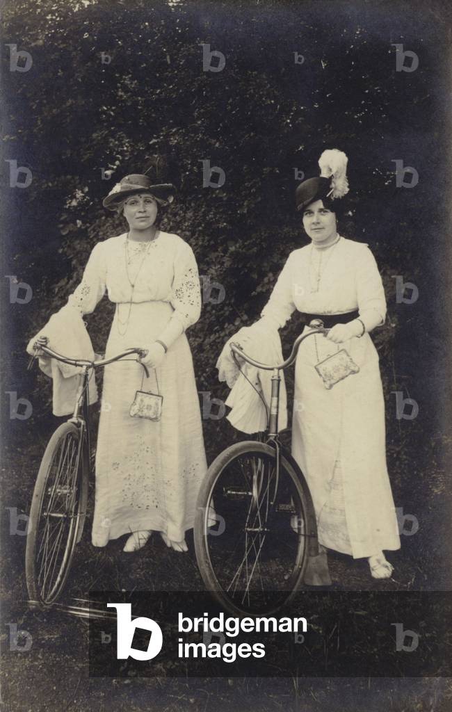 Two women with bicycles, carrying purses (b/w photo)