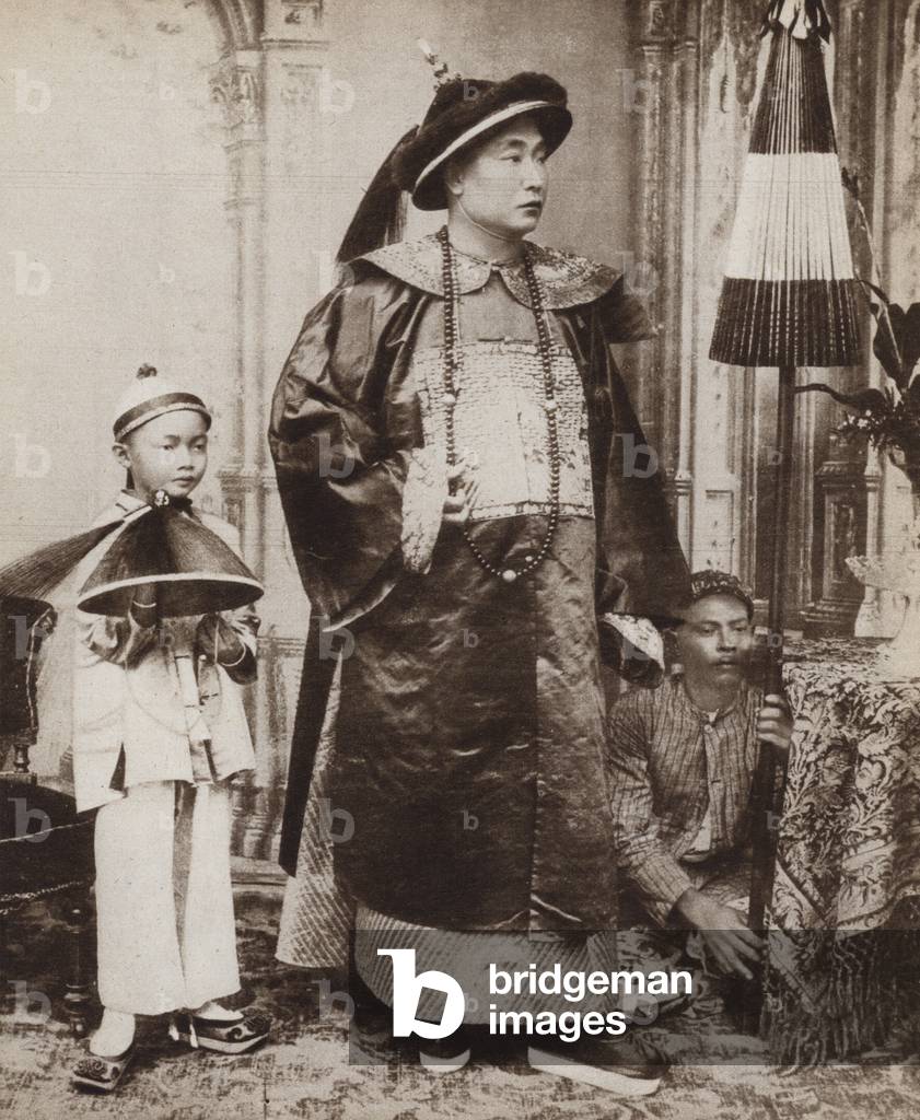 Chinese military mandarin wearing traditional costume with his servants holding his headdress and umbrella (b/w photo)