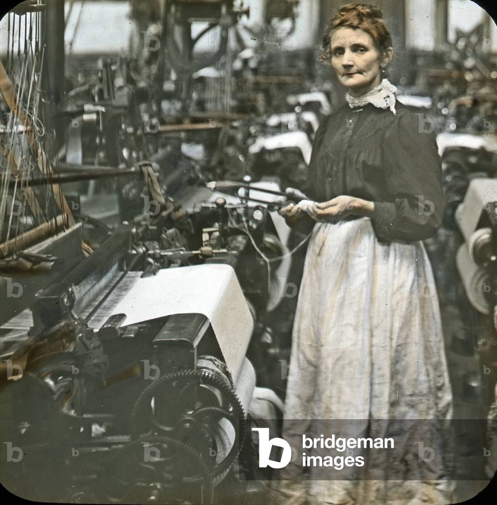 Weaving cotton: woman in white pinafore stands at a loom in a textile mill, probably Leeds, 1905-15 (colour lantern slide)