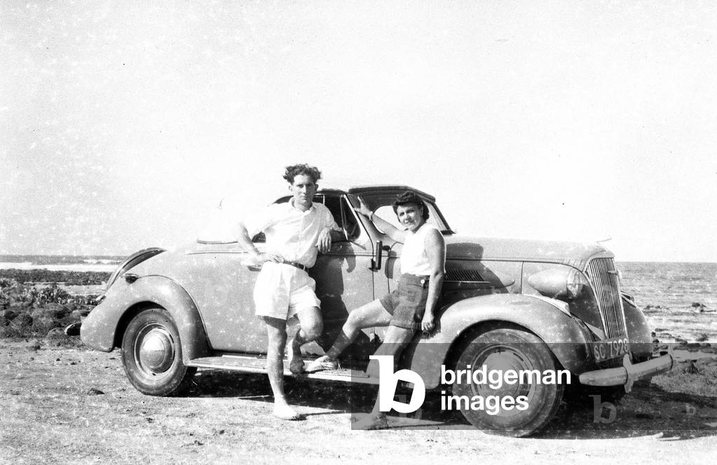 Africa: Decapotable car in the African bush with young couple sitting on the hood, 1947 -