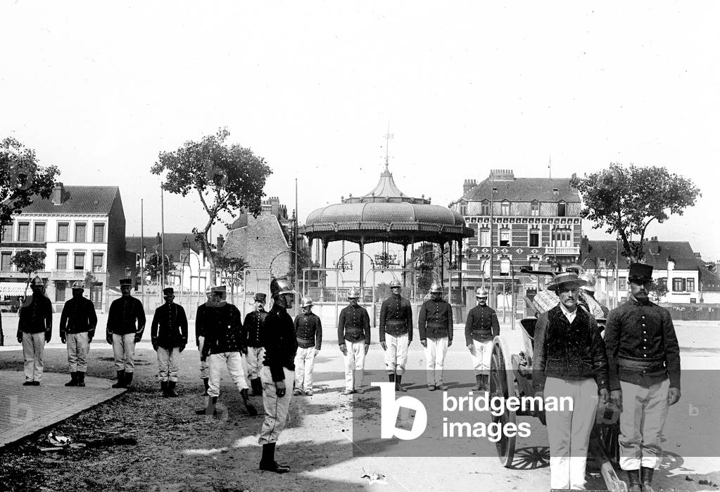 France, Nord-Pas-de-Calais, Nord (59), Roubaix: fire brigade exercises, firefighters with their helmet arrive running on the square of a church under construction pulling a hand pump. Presentation of firefighters and fire pump, lined up at the foot of a kiosk on church square, 1890