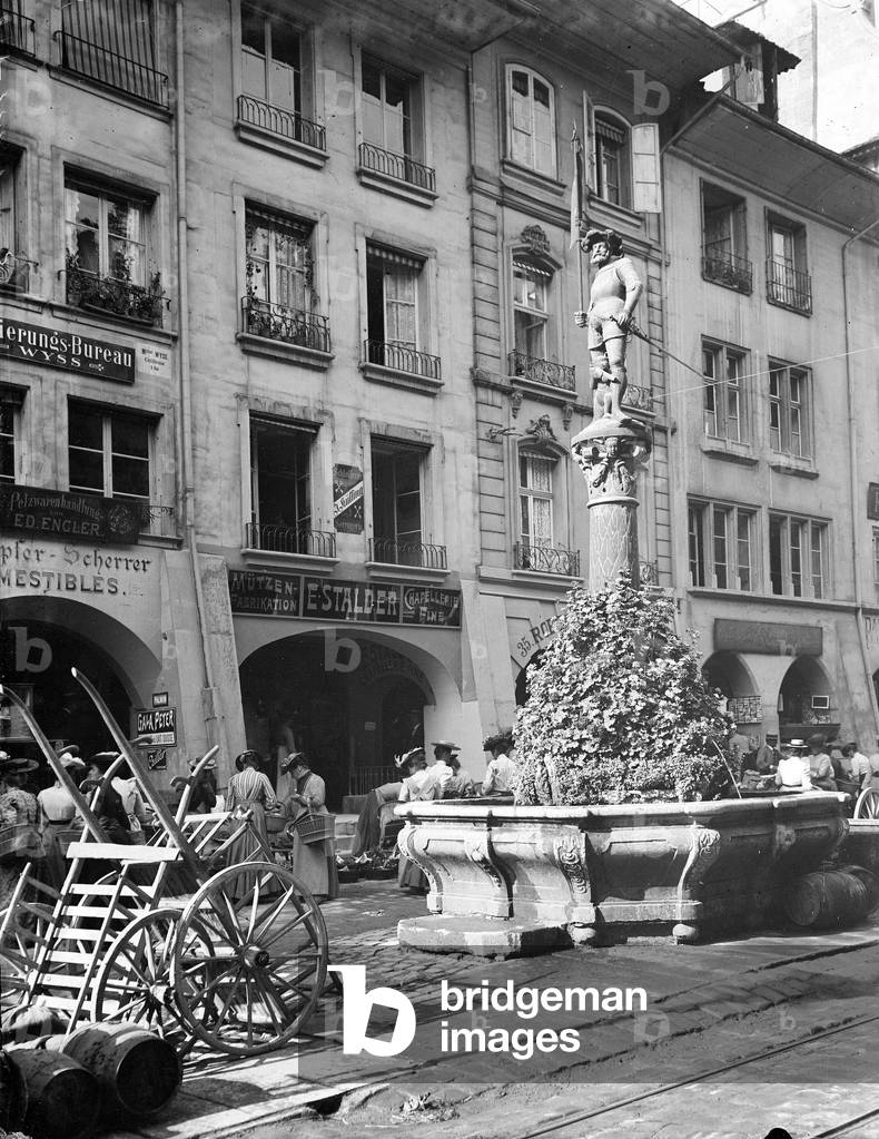Switzerland, Canton of Bern, Bern: the Marktgasse on a day walk with a fountain representing a banner holder with between his legs a small bear wearing armor and firing a gun shot, 1895