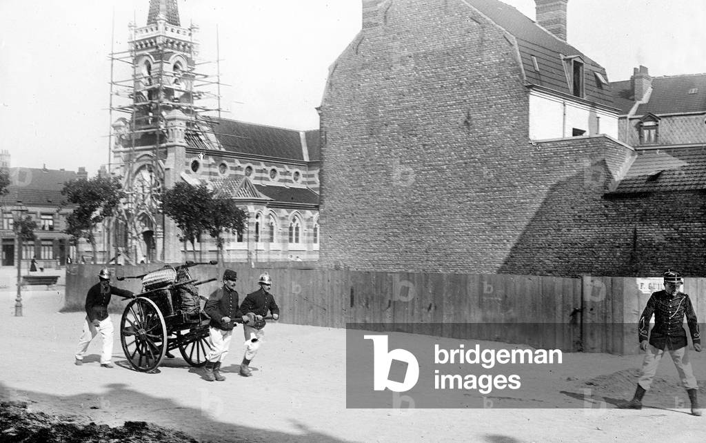 France, Nord-Pas-de-Calais, Nord (59), Roubaix: fire brigade exercises, firefighters with their helmet arrive running on the square of a church under construction pulling a hand pump, 1890