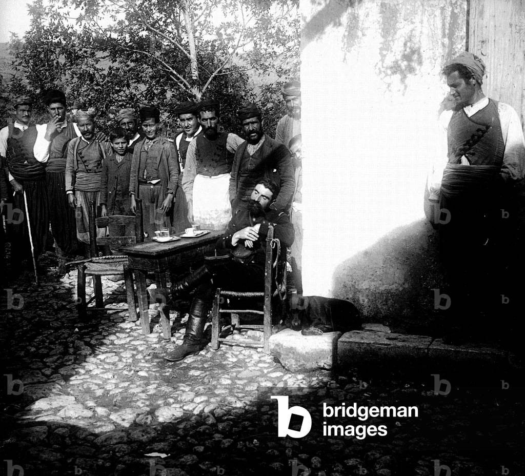 Greece, Crete, CR, Hierapetra, Ierapetra: French sector, terrace of a cafe with many indigenous people sitting next to a French soldier sleeping - Small table and chairs on a pave courtyard, 1906