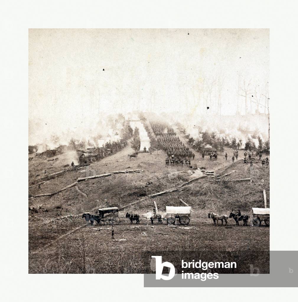 American Civil War: Three Horse-Drawn Covered Wagons in the Foreground. Soldiers Marching in Formation Between Rows of Small Cabins and Tents in the Background. Photo, Albumen Print