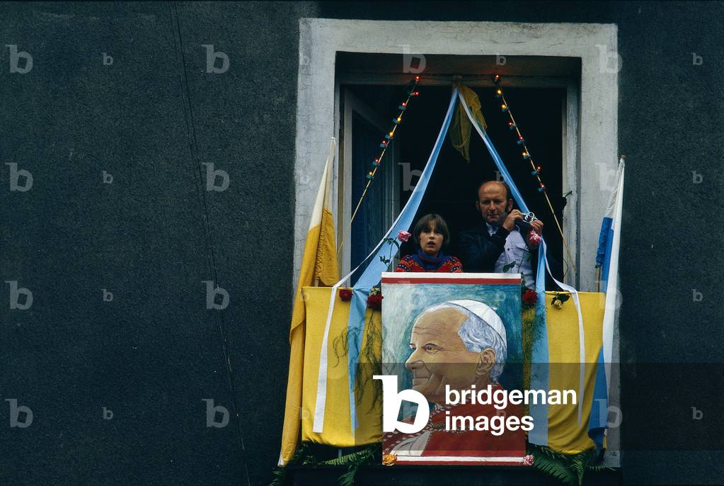 Two people are appearing at a window decorated with a portrait of the pope, on the occasion of Pope John Paul II to Poland
