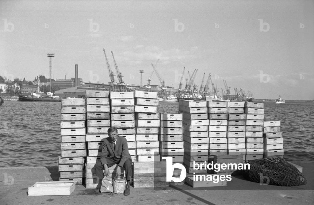 A man posing seated on wooden crates, Behind him, the catch of the day, Finland, 1960s (b/w photo)