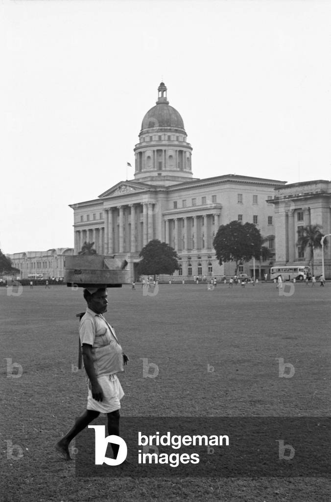 Man carrying items on the head at the park nearby the Old Supreme Court of Singapore, Singapore, 1962 (b/w photo)