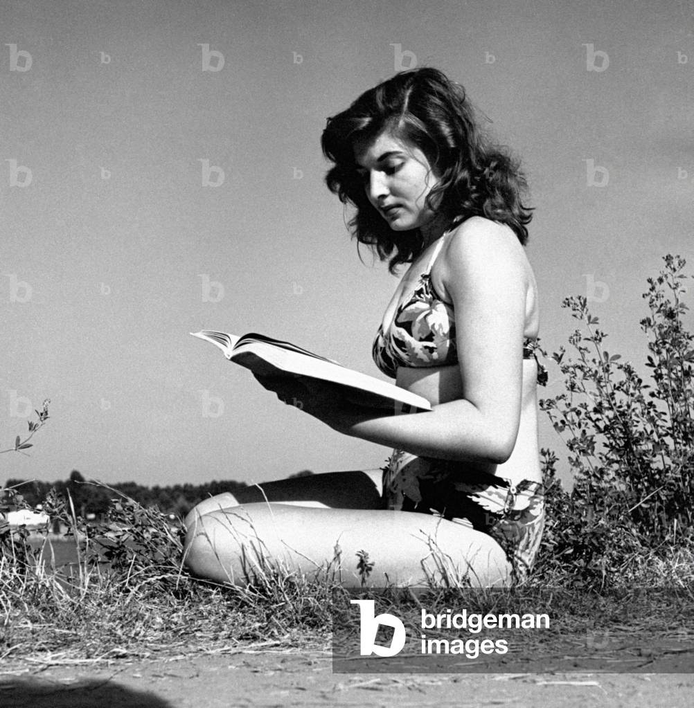 A girl reads a book sitting on the grass, Italy, 1955 (b/w photo)