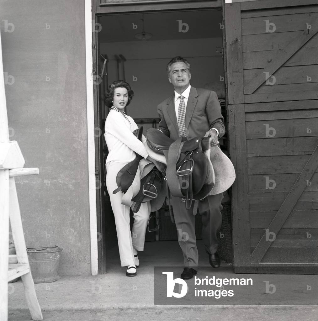 Rossano Brazzi and Dawn Addams holding two saddles, Italy, 1957 (b/w photo)
