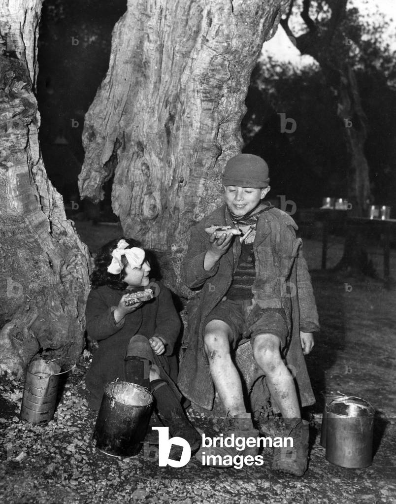 Two Italian children eat at the foot of a tree