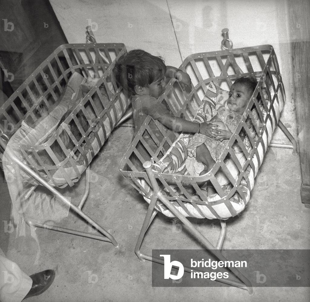An ill baby in his cradle is being caressed by a child at the Salesian community of Fr. Orfeo Mantovani, 1966 (b/w photo)