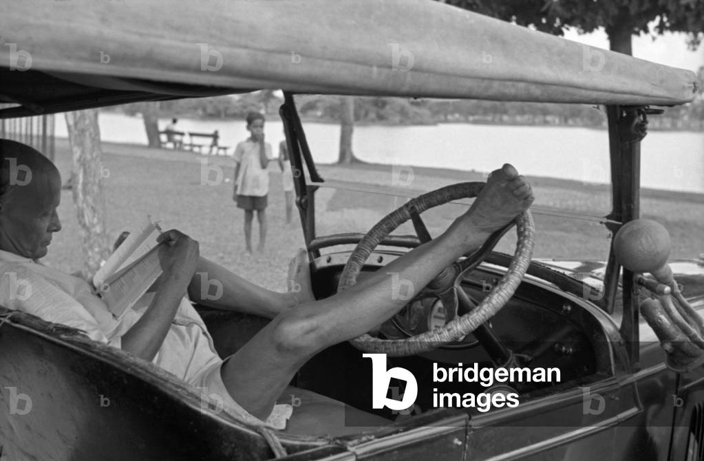 A man reading a book on a car, Kolkata, India, 1962 (b/w photo)