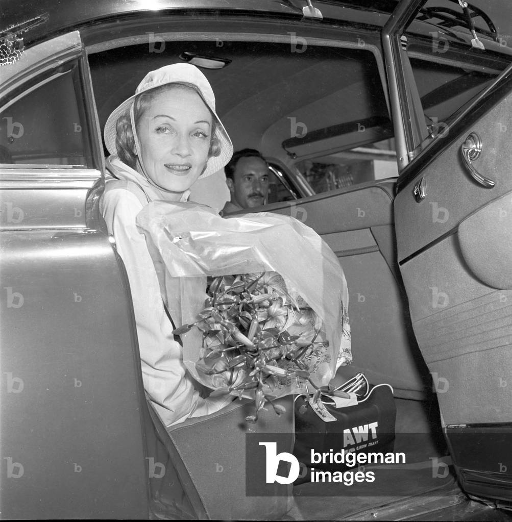 Marlene Dietrich getting into a car with a big bunch of flowers, 1957 (b/w photo)