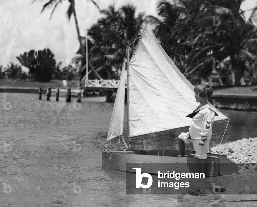 A child stands on the shore by a hand-made, one-person sailboat, c.1930 (b/w photo)