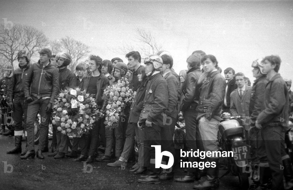 Ton Up Boys Funeral:
The mourning dress was black leather.
Motorcycles were parked as eighty youngsters solemnly flanked the half mile approach road to the crematorium, to watch the cortege go by.
Then sadly walked behind to the service.
The coffee bar set were honouring a promise made with their colleague Michael Munnerley, 18 killed in an accident on his bike.
A pact that promised if any of them died the rest would ride to his funeral on their bikes. 
Our Picture Shows: Motorcyclist seen here outside the crematorium before the funeral. January 1969
Z1129