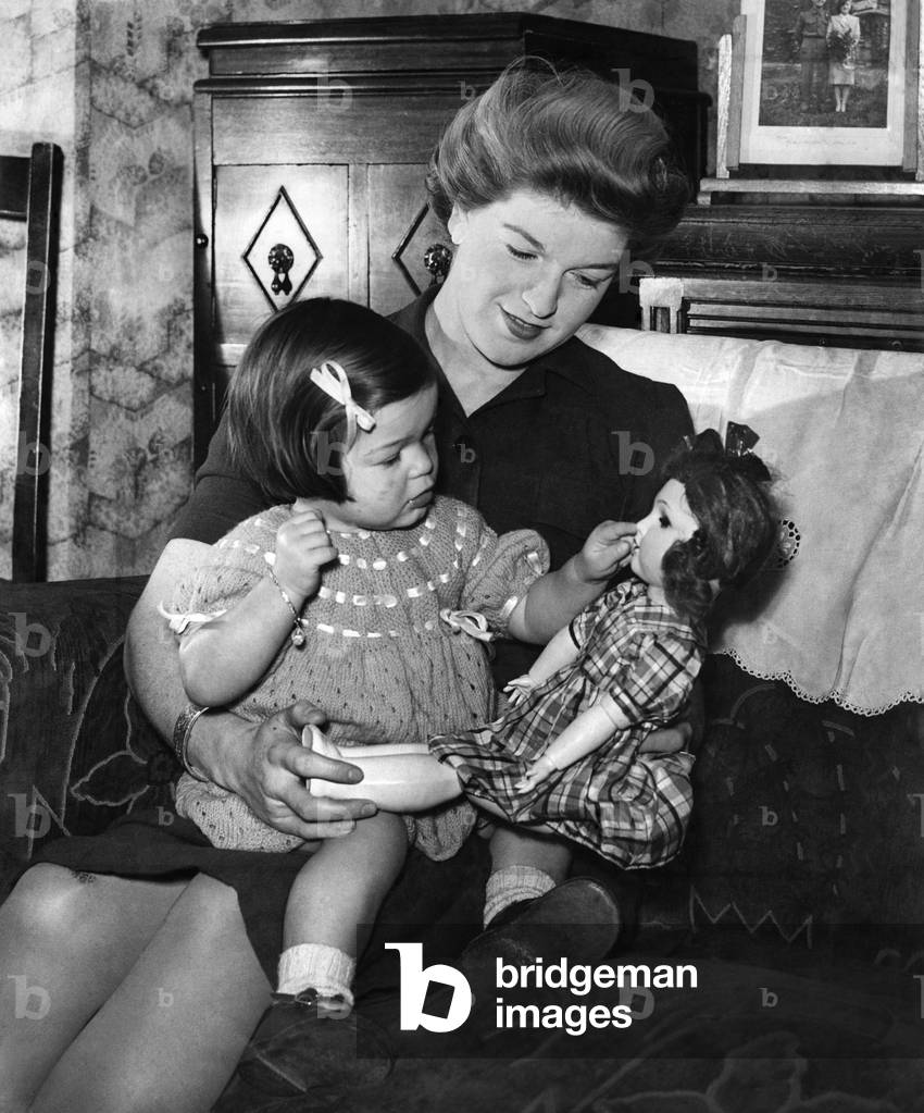 Mother and child at home, the girl playing with her doll as she sits on mummy's lap. 
Circa 1942