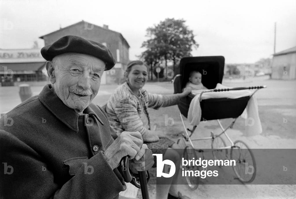 An old Frenchman sits out in the sunshine, near Toulouse, in the background is a mum and baby, April 1975 (b/w photo)