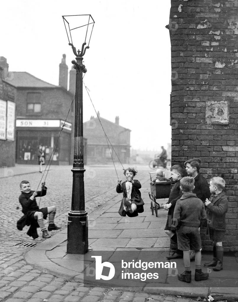 Children in a Manchester Street find their own enjoyment with the aid of a rope and a lamp post. Children playing in the street, swinging from a streetlight in March 1943 (b/w photo)