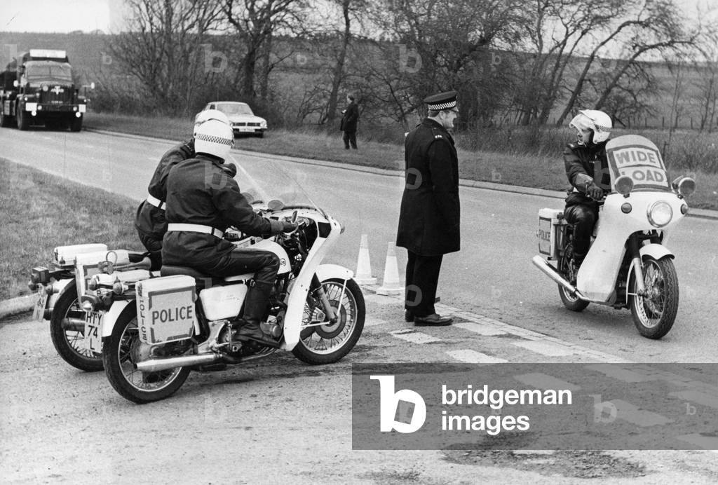 Police on motorcycles at the junction of a road easing the passage of armoured vehicle, c.1970