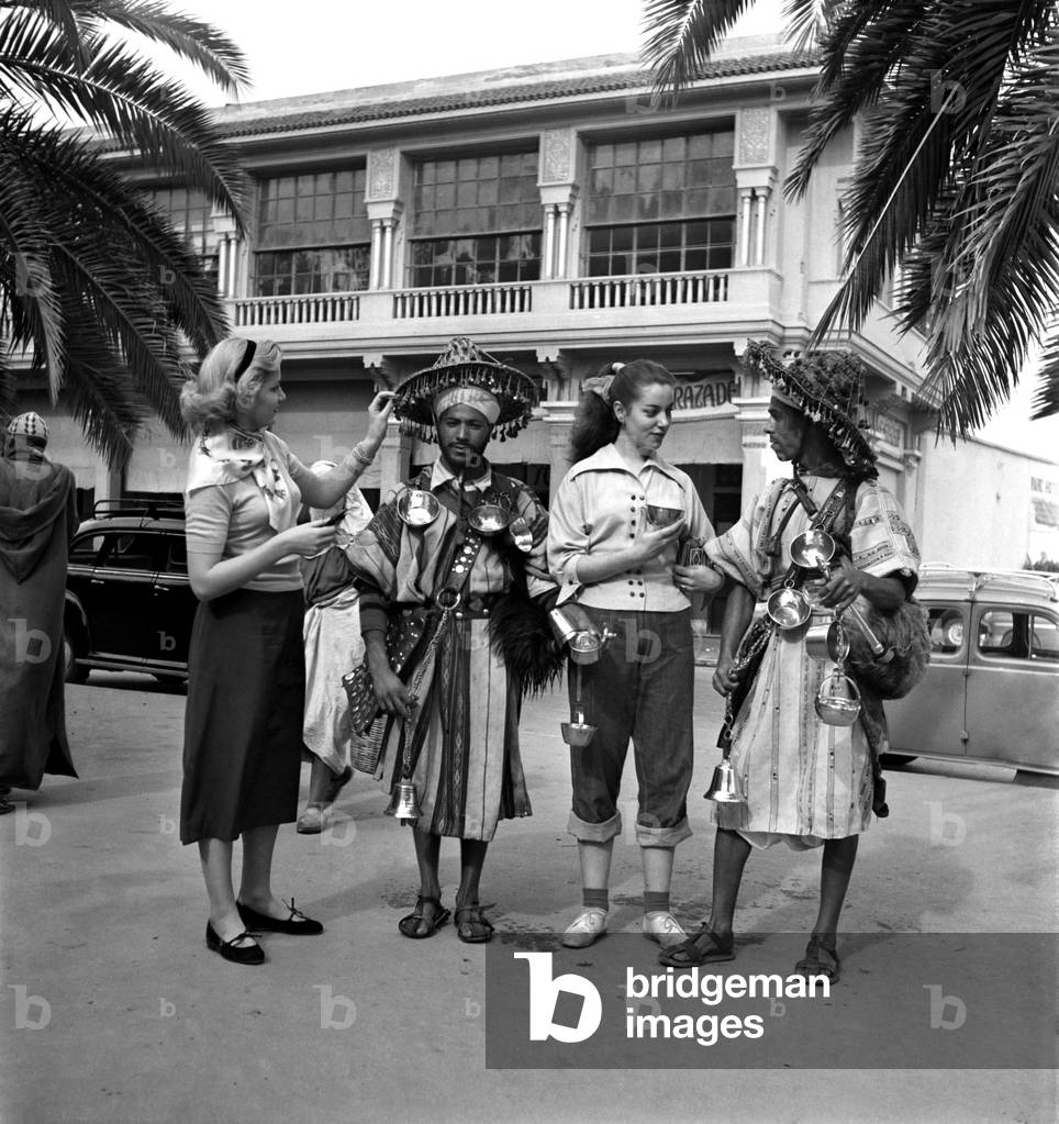 French actresses buy a drink of water from a local vendor in the 
town of Marrakech during their visit to Morocco. December 1952 C5919-002