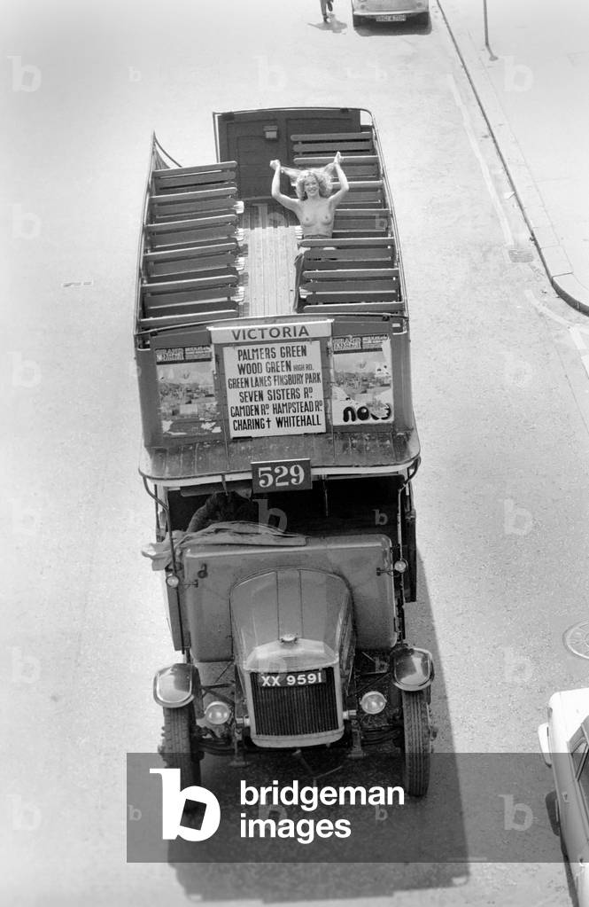 Topless model Sarah Walkden sitting on the top deck of an open top London bus as she takes a tour of the city, May 1975 (b/w photo)