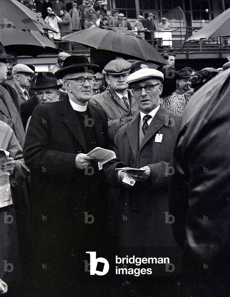 A priest holds his guide during a day out at the races in Cheltenham, March 1963 (b/w photo)