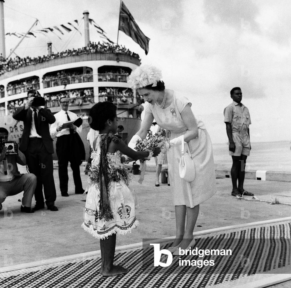 Queen Elizabeth II arrives in Suva, Fiji from the Royal yacht and is presented a bouquet of flowers by Fijian girl Adi Kaunilotuma who sat down in front of her on the carpet during the royal visit to Fiji, February 1963 (b/w photo)