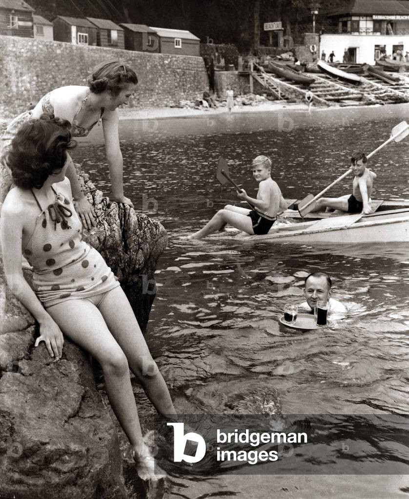 A family sitting on the rocks and playing in the water at Babbacombe Bay in Devon as a waiter swims out from the bar to bring over drinks for them, 11/09/1948 (b/w photo)