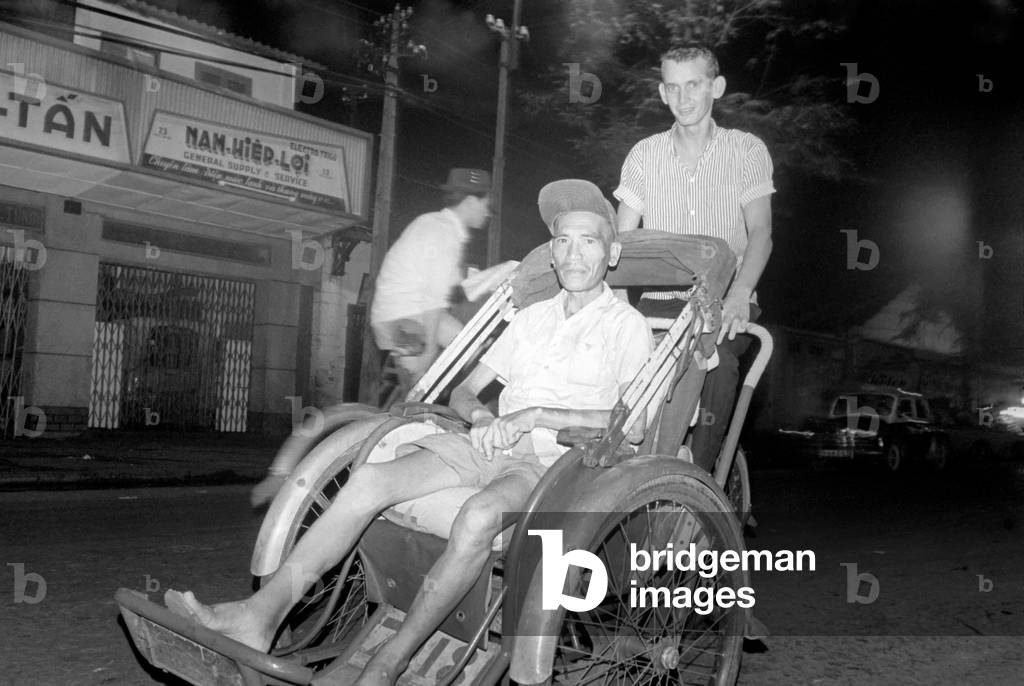 Night life in Saigon Vietnam. Young man pushes another man in a type of rickshaw along the street, October 1966 (b/w photo)
