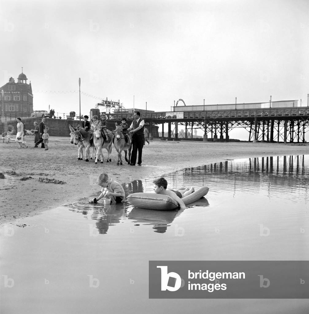 Fun on the beach at New Brighton, Cheshire, for three-year-old David Pearson, from Birkenhead, aboard his dinky dinghy. Fun for Carl Tharme, 9, of Liverpool, who preferred a paddle. Others take a donkey ride near the pier, March 1961 (b/w photo)