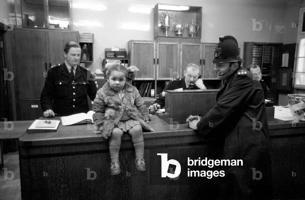 Police and Children. The scene is set in the enquiry office of Bishopsgate Police Station, in London, where to-day in the street nearby the Petticoat Lane thousands of shoppers were busy trying to find bargains. Seated on the counter is a little girl brought in by Police Constable B 232, who had apparently lost her mother in the crowd. December 1969