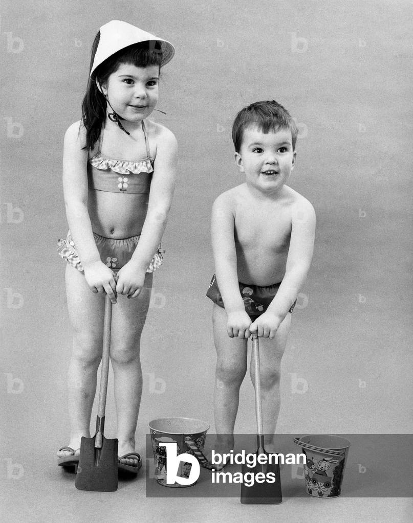 Fashion 1960s.
Children play with bucket and spade in their swimwear, a little bikini and hat, and pair of trunks.