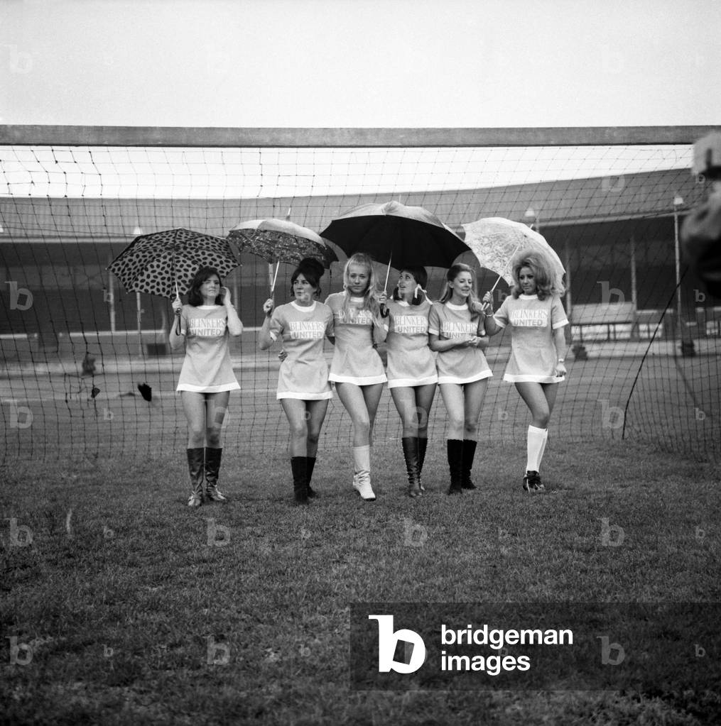 Lined up in their mini soccer dresses at what should have been the time for kick-off are 6 of the Blinkers Utd football team. The Blinkers Utd v All Stars XI charity football match had been cancelled after it was decided that the weather was not fit to play. As the men watched the girls ran around the ground at Manchester's Belle Vue carrying umbrellas to protect their hair. November 1969 (photo)