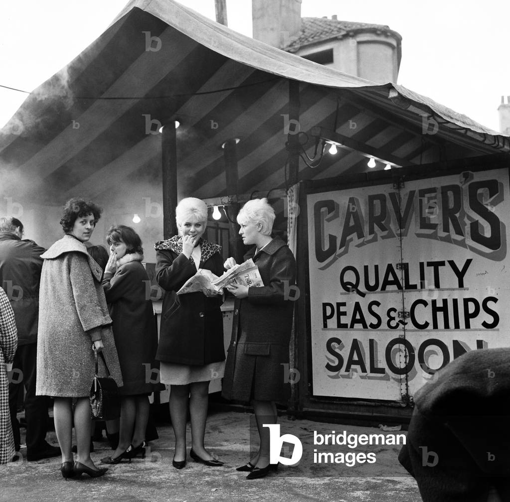 Jeanne Stather (22) and Jennifer Fussey (19) enjoy lunch time chips from the open air chip shop at the market. Hull, East Yorkshire. 16th March 1965 (b/w photo)
