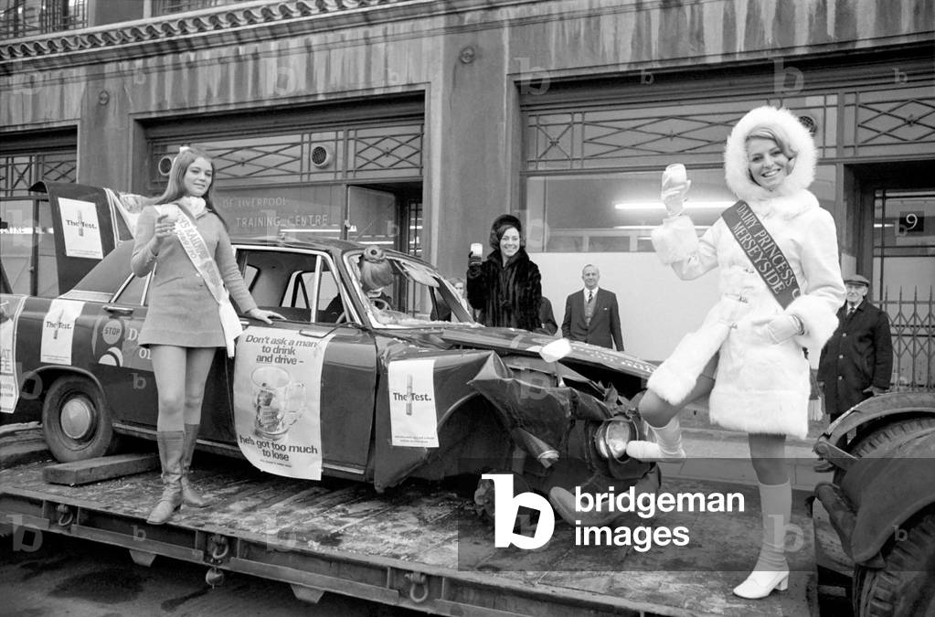 As part of Liverpool's keep death off the roads campaign this Christmas a low loader is touring the city with a smashed up car aboard. To give the campaign a send-off, two beauty queens and a personality girl drank a toast in milk to wish the campaign success. The beauties were the National Dairy Queen, Miss Gaynor Lacey, 20, the National Railway Queen of British Rail, Miss Marilyn Pope, 18, and Miss Littlewood, Miss Ida Bailey, 24. December 1969