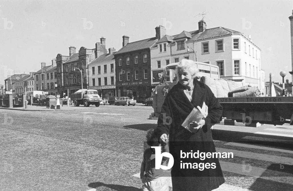 A grandmother and her grandaughter crossing the main road in Lurgan after paying a visit to the newsagent. Circa 1966