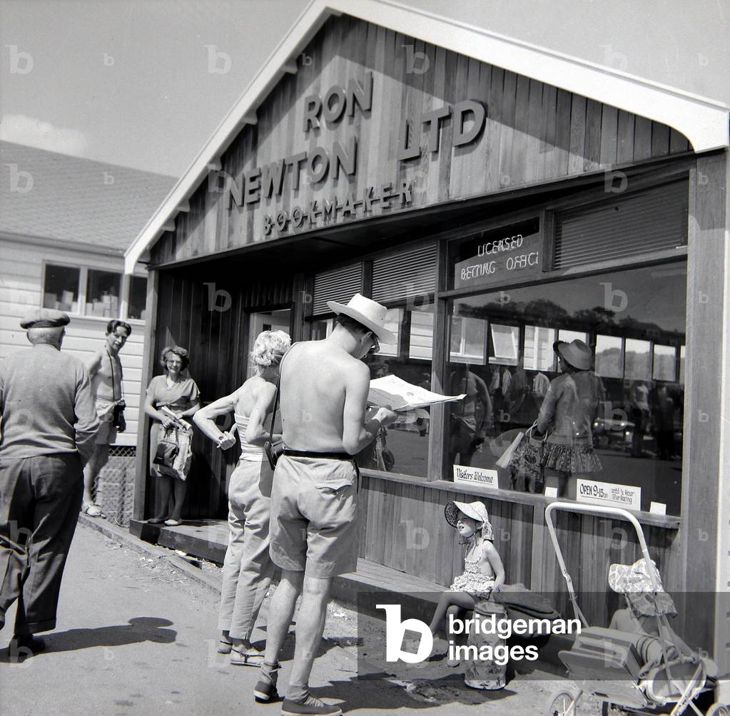 Holidaymakers study the form guide at a betting shop in the form of a wooden chalet on the beach at Dawlish Warren in Devon
July 1962