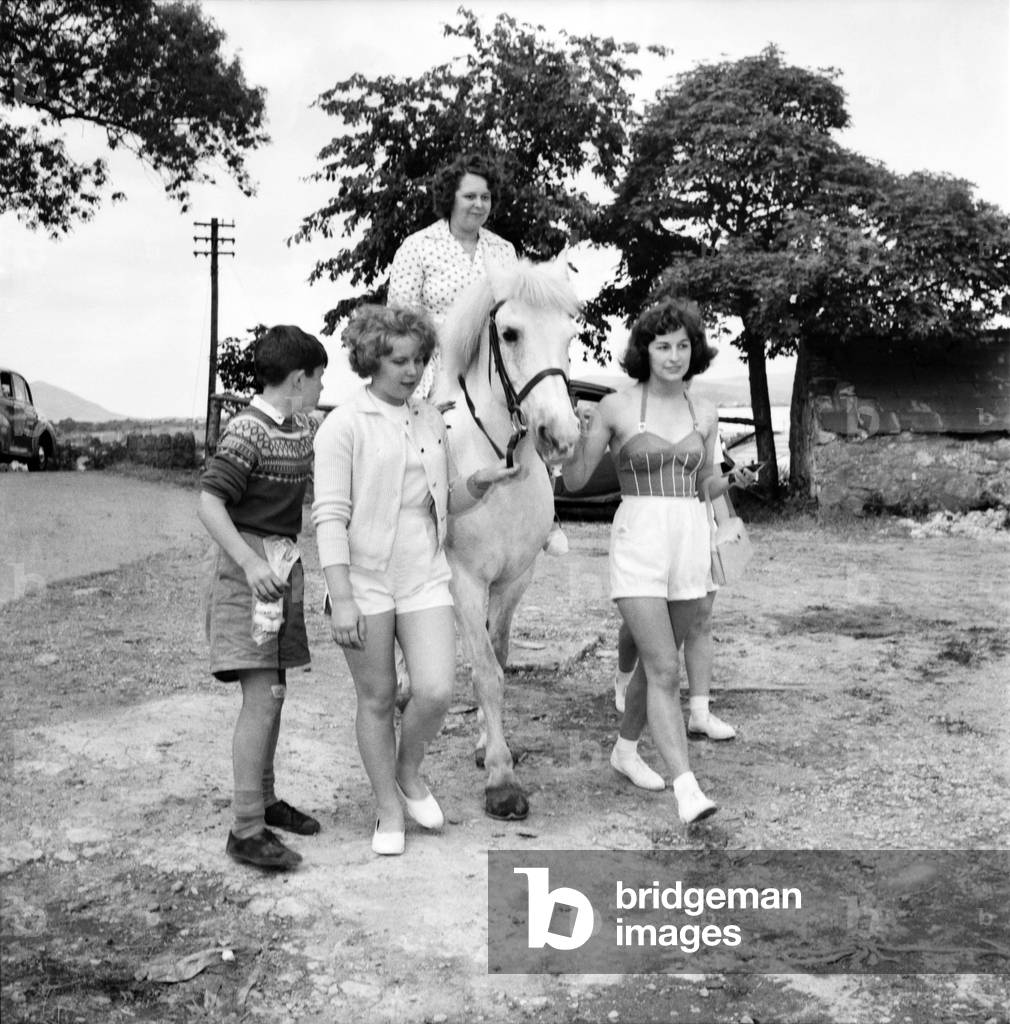Bultins Holiday Pwllheli: Mrs. Nellie McGrail, Pools Winner. Horse riding/Boating/Elephant/Roller skating/Family. Mrs. McGrail is helped into the saddle of a camp horse by daughter Irene. Daughter Barbara holds the horse's head (left) with Brenda on right. June 1960