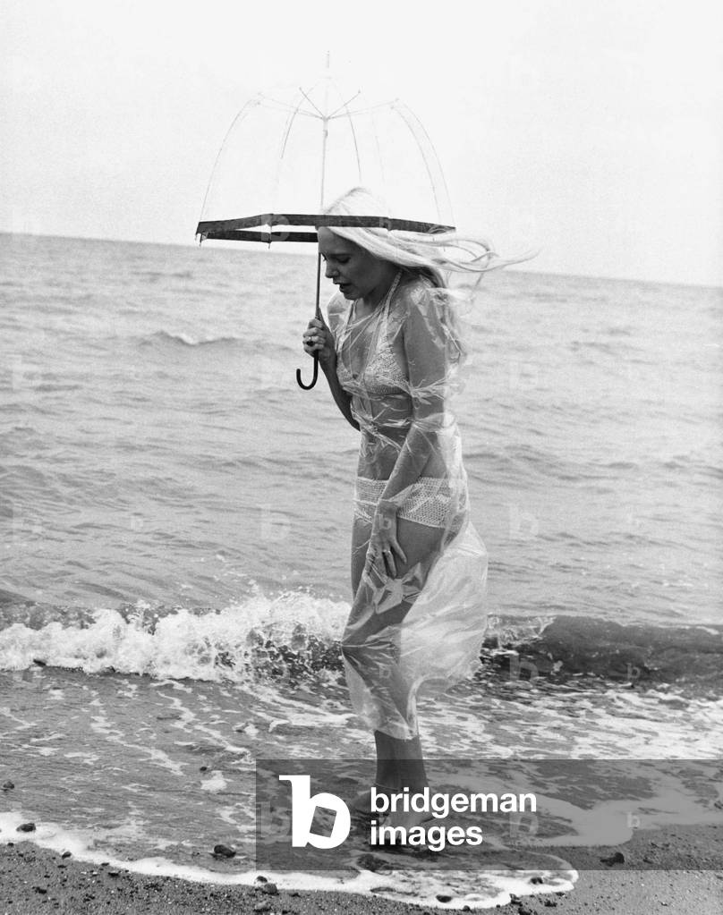 Seaside Beaches Beach Scenes. Model Flanagan takes a walk / stroll along the beach wearing bikini and clear plastic bag to protect herself from the biting wind, June 1972 (b/w photo)