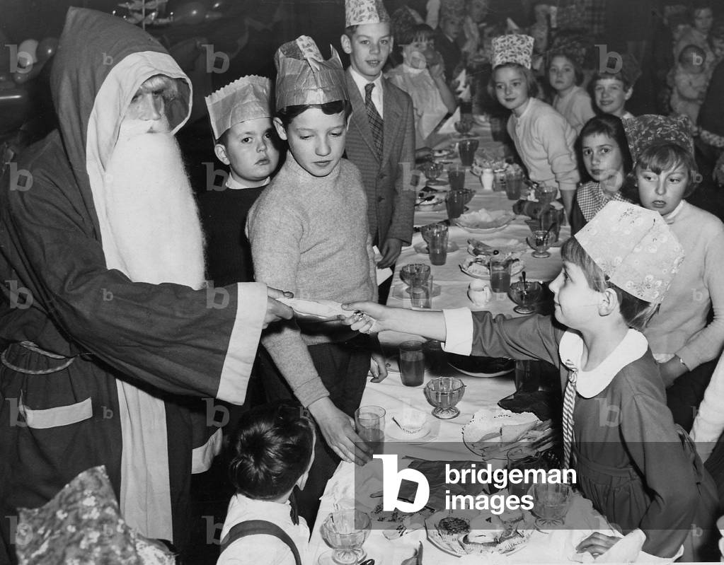 Father Christmas, arriving with a present for each of the 70 children, helps seven-year-old Susan Stevens, of Castle Bromwich, to pull a cracker at the party given in Kyrie Hall by the garage staff of The Birmingham Post & Mail Ltd, 03/12/1960 (b/w photo)