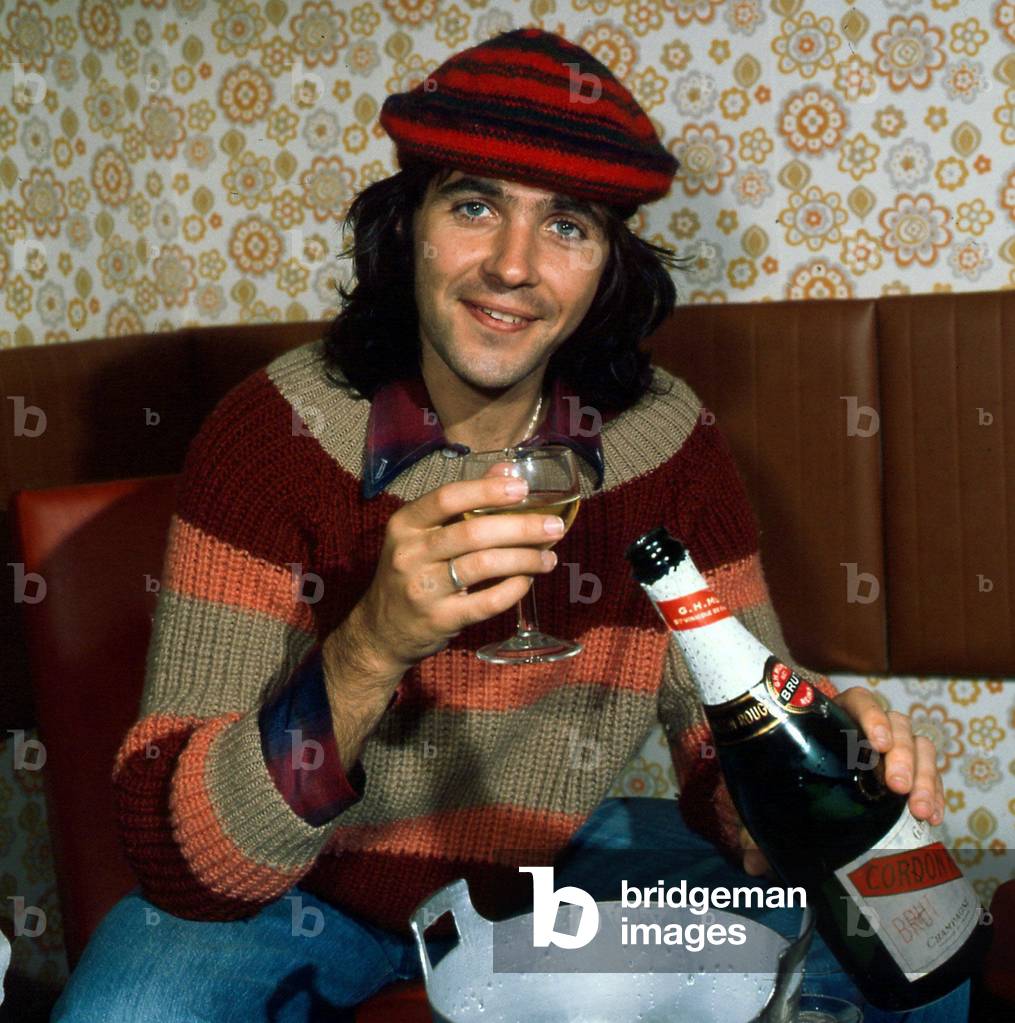 A portrait of David Essex, taken in Scotland, wearing a multicoloured pullover and a tammy hat, holding a bottle of Champagne, 24th September 1975 (photo)