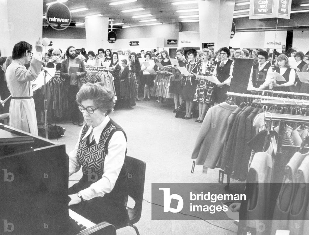 All is safely gathered in... Staff at Own Owen, Broadgate, Coventry, singing hymns before the store opened. It is the second year running that a harvest service has been held at the store. This year, the Coventry Cathedral choirboys sang at the service. Staff brought produce which was later distributed to the elderly in the city. Perishable goods were being given to pensioners who had worked at Owen Owen, at their monthly tea party today. The Rev. Denis Claringbull, the industrial chaplain, and the Rev. Nigel Abbott, Vicar of Holy Trinity Church, conducted the service, 8th October 1974 (b/w photo)