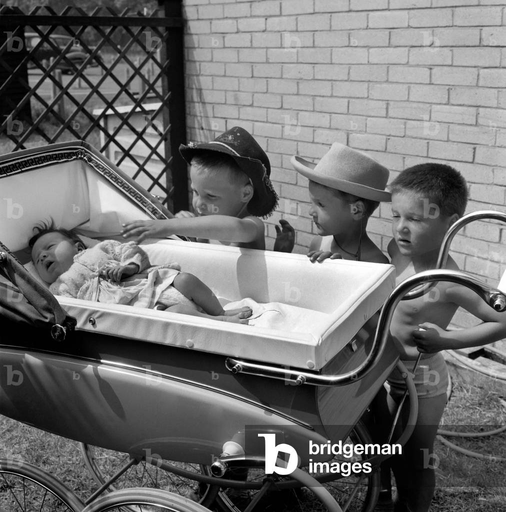 The three gloomy-faced triplet brothers all wearing cowboy hats leaning over the side of a pram in the garden of their home at their new born baby sister. 
June 1960