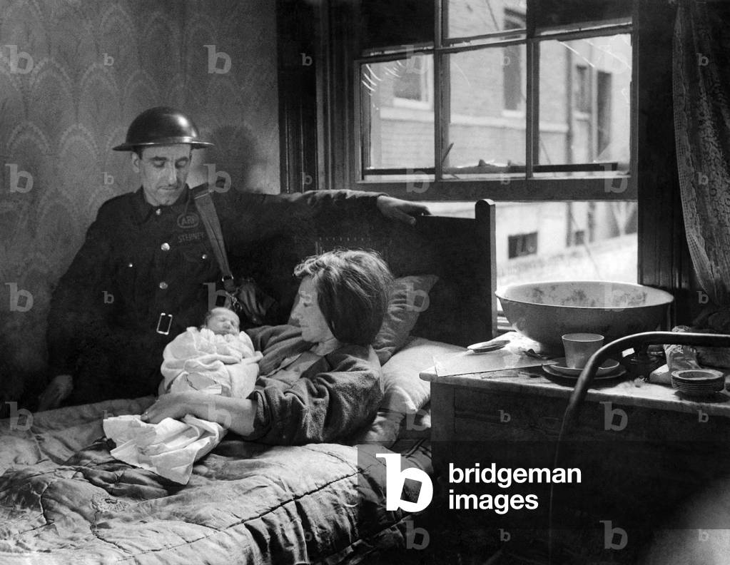 Warden Bill Watson at the bedside of Mrs. R. Foster. With bombs falling
round, as Mrs. Foster lay on that bed in darkness, and shattered glass, Mr.
Watson acted as midwife at the unexpected birth of her child during the first large scale air raid on London. 8th September 1940